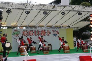 「おおい祭り」と書かれた舞台の上で、和太鼓が並べられており、和太鼓の前に一人ずつ立ち、バチを持って右手を上げ、演奏をしている様子の写真