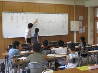 前方の黒板を使って講師の先生が説明をしており、その説明を教室の子ども達が真剣に話を聞いている写真