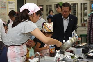 大人の女性が鍋を持って小さい器にうつしており、エプロンと三角巾を身に着けた子どもたちが調理台の前で作業をしているのを横から見ている様子の写真