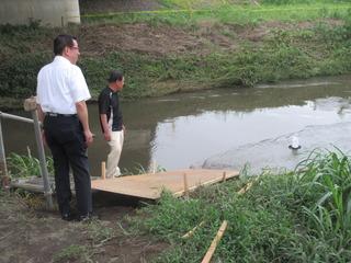 河川敷から川に向かっ木の橋が作られており、一つの灯籠が流されている様子を市長と白いズボンをはいた男性が見つめている写真