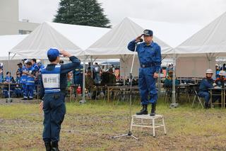 市長が訓練表の青い服を着て台の上に立って、進行班のゼッケンをつけた男性と向かい合って、右手を顔の横に当てて、敬礼をしている写真