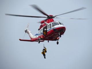 赤と白色のヘリコプターが上空を飛んでおり、ヘリコプターからロープにつながった人が下がっている訓練の様子の写真