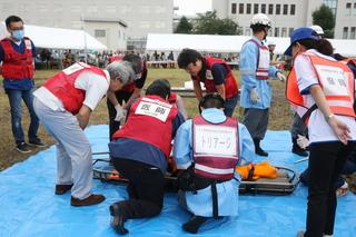 ブルーシートの上に担架が置かれその周りに医師や トリアージのベストを着けた人が集まっており、訓練が行われている様子の写真