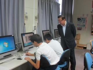 パソコンの並んでいる教室で男子生徒が、パソコンの前で学習しており、その様子を後ろから市長が見ている写真
