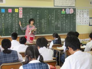 先生が黒板に板書した文字を指差しながら説明をしており、生徒が席に着いて授業を受けている写真