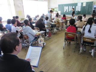 教室にて、先生が教壇に立って授業をしており、市長が教室の子ども達の席の後ろに座って授業風景を見ている写真