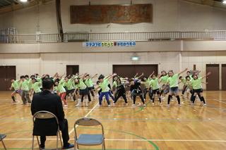 黄緑色のお揃いのティーシャツを着た3年生の子ども達が左手を上に挙げて、右手を下に下ろした同じ振りでダンスを踊っており、市長がパイプ椅子に座ってダンスを見ている写真