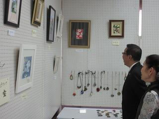 壁に額に入った絵画や、手作りネックレスが展示されており、市長と女性が作品を見ている写真