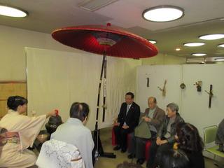赤い大きな日本傘の下で茶会が行われており、茶席に座っている市長が着物を着た女性のお茶のお手前を見ている写真