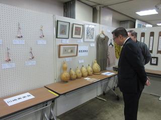 展覧会の作品展で、机の上にひょうたんが並んでおり、壁には手作りの作品や絵画が飾られており、市長が作品を見ている写真