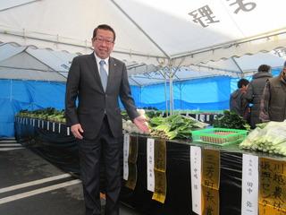 JAいるま野秋の収穫祭農産物品評会にて市長がテントの中の机の上に並べられている野菜の横に立ち手の平を向け紹介している写真