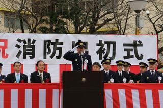 紅白の横断幕が長机の周りに巻かれており、壇上yに立っている消防の制服を着た市長が右手を顔の横に付け敬礼をしている写真消防出初式1