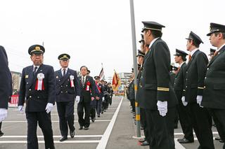 消防の制服を着て、胸に白い胸章を付けた市長が、関係者の方々と1列に並んで行進をしている写真