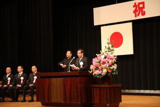 日の丸の国旗が壇上の幕に掲げられており、きれいな壇上花の横で、市長が祝辞を述べており、市長の横で手話の同時通訳者の男性が通訳をしている写真