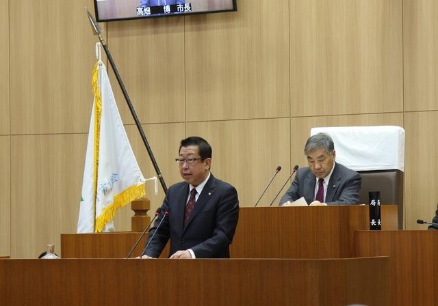 議場で演壇に立ち話をしている市長とその後ろに座っている議長の写真