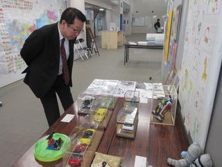 テーブルの上に展示されている透明な箱の中の車やロボット等のプラモデルを鑑賞する市長の写真
