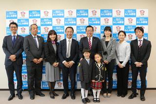 新小学1年生の男の子と女の子が贈呈された黄色いワッペンをつけて贈呈業者や市長たちと記念撮影している写真