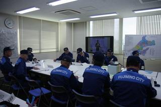 総合防災訓練にて、会議室に関係者の方々が向かい合って座っており、電話対応をしていたり、机の上の書類に目を通したりしている訓練の写真