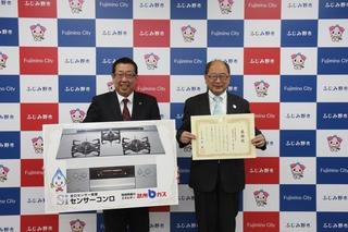 武州瓦斯株式会社の代表者が感謝状を持ち、市長がセンサーコンロの写真のパネルを持って立っている写真
