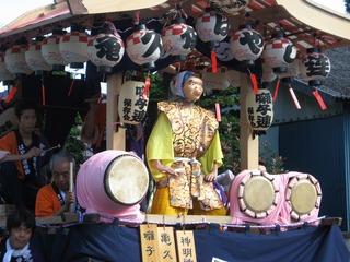 木組みの屋台の上で笛と太鼓と鉦による賑やかな演奏の中ヒョットコの面をつけた男が踊っている写真