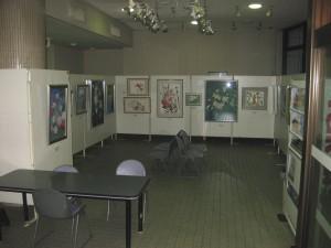中央に背中合わせに並べられた4台の椅子と、それを囲むように設置されたパーティションに絵画がかけられているギャラリーの写真