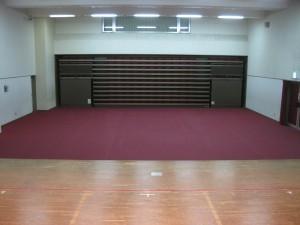 赤紫色の床があり、正面奥に電動収納椅子と左側に出入り口があるホールを、手前のステージの上から撮影した写真
