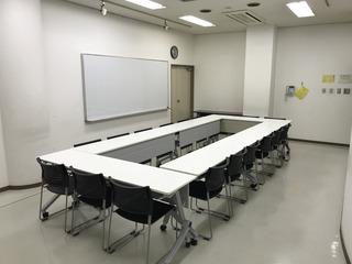 壁にホワイトボードが設置され、長方形に設置された長机の奥に出入り口の扉がある第1学習室を、部屋の隅から撮影した写真