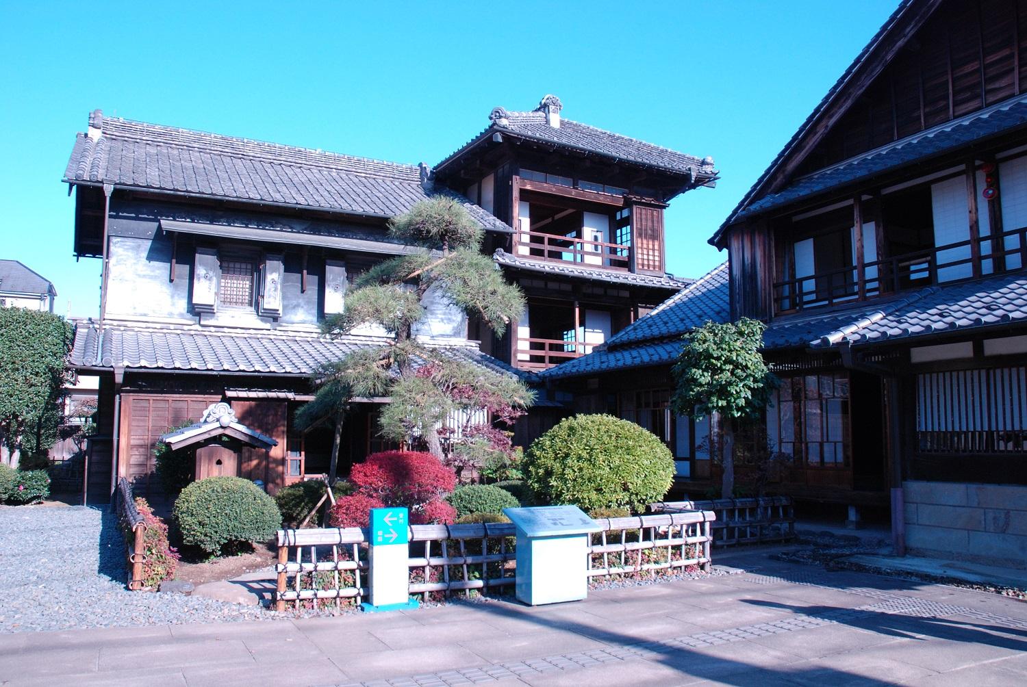 青い空を背景に、松の木と赤や緑の庭木が植えられたお庭とその奥に建つ3階建て古民家の写真