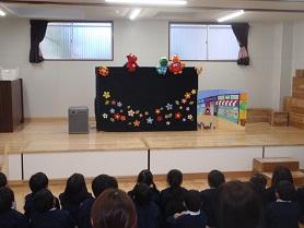 舞台の上で行われている怪獣の人形劇を座って見ている幼稚園児の写真