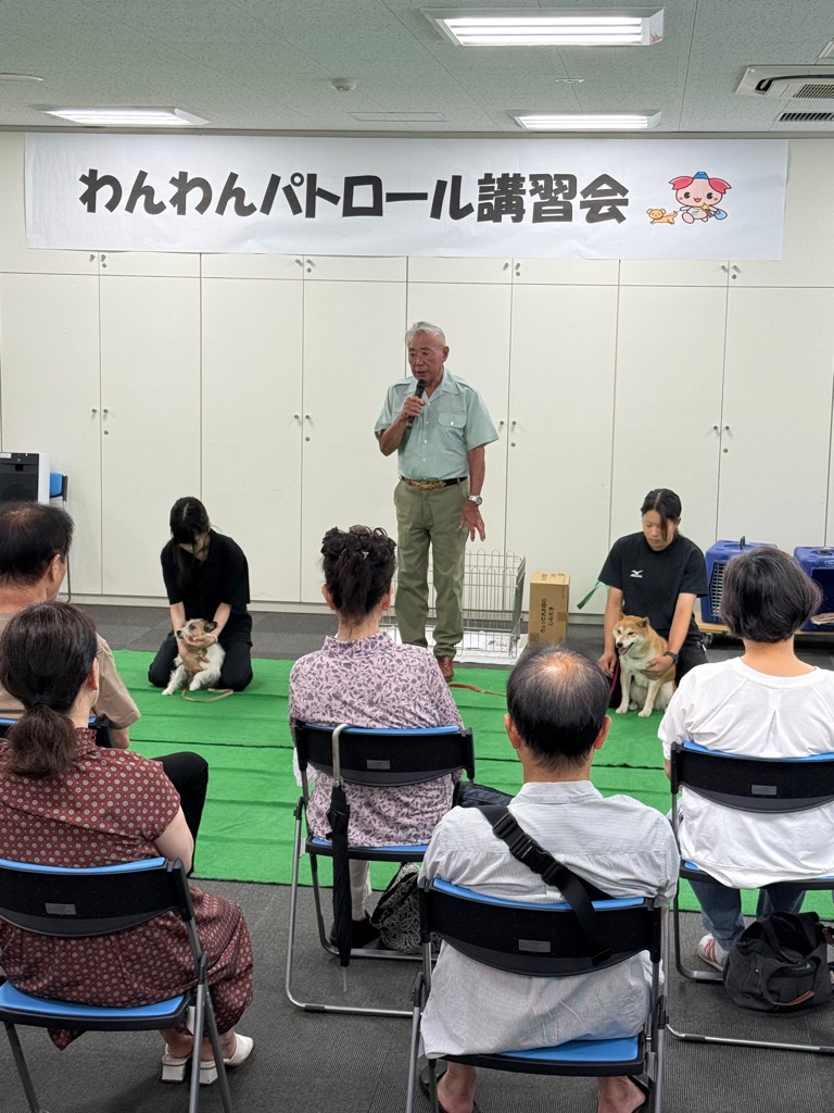 座っている講習会参加者の前にマイクを持った講師、実演者2人と犬2匹がおり、説明を行っている写真